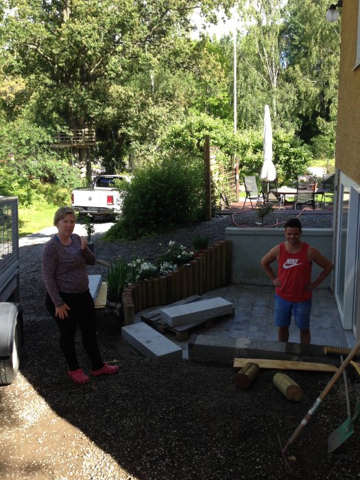 Två personer står vid en trädgård under renovering med granitstenar och verktyg synliga.