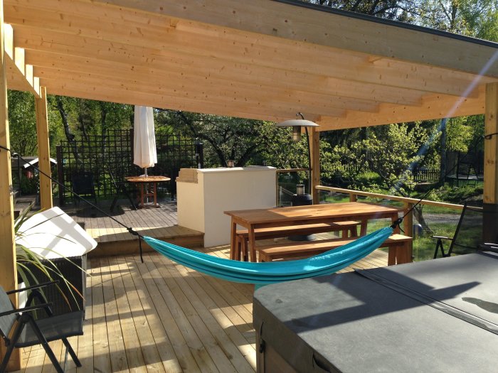Träaltan med tak, hängmatta, bord och bänkar i soligt trädgårdsmiljö.
