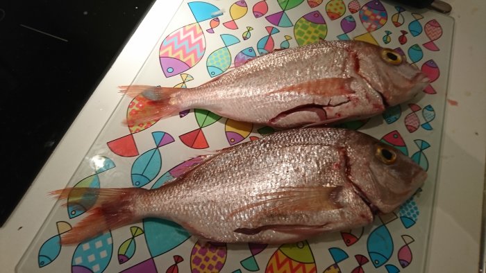 Två hela fiskar med rosaorange fenor på en skärbräda med färgglada fiskmotiv.