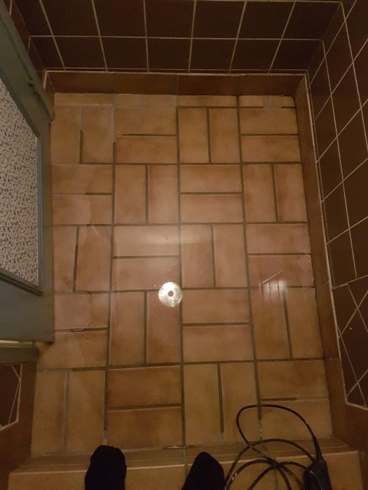 Tegelklädd trappa med vatten och avloppshål, synliga vattenspår och personens fötter.