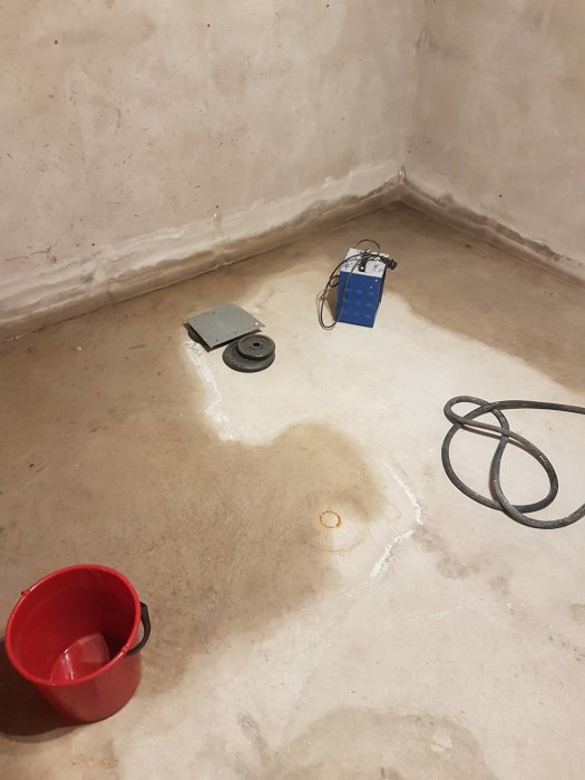 Källarförråd med vattenläckage och dräneringspump, röd hink och borrat hål i betongsulan.