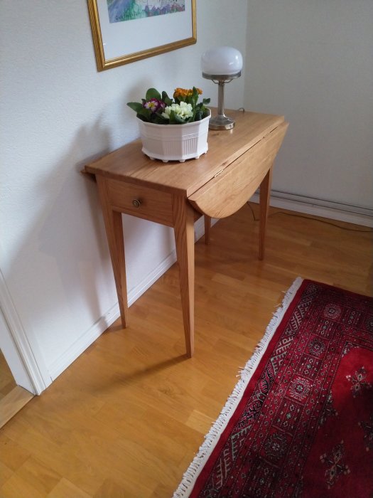 Träbord med låda, blomkruka och lampa i ett rum med orientalisk matta och tavla.