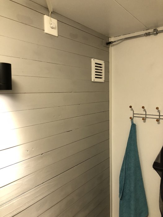 Ett hörn av ett badrum med fuktspår på en grå panelvägg, en handdukshängare och en ventilkåpa högt upp på väggen.