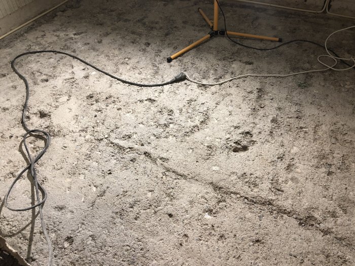 Grov betongplatta i källarplan med synliga repor och fördjupningar, samt två gula verktyg och elsladdar på golvet.
