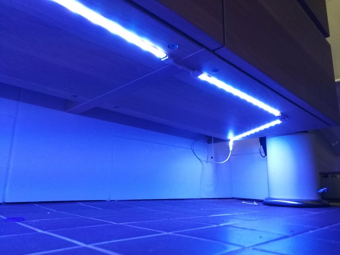 LED-strip monterad under köksskåp som lyser med blått ljus, kablar synliga, på mörkt golv.