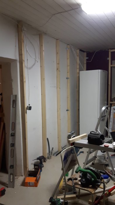 Renoveringsprojekt med nyuppsatta reglar på en vägg, verktyg och stege i förgrunden och kylskåp i bakgrunden.