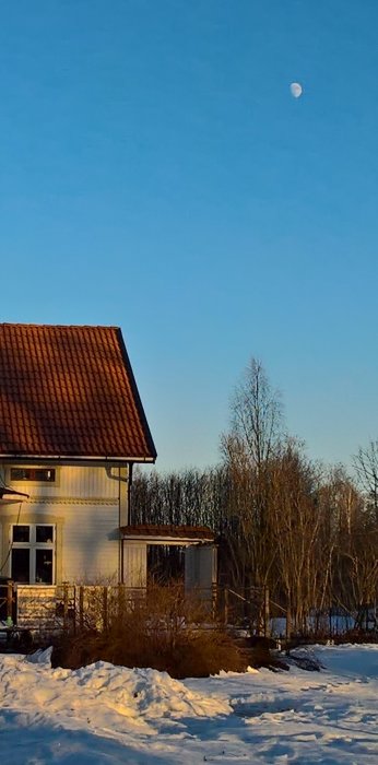 Husfasad med takpannor mot en klarblå himmel med synlig halvmåne och snötäckt mark.