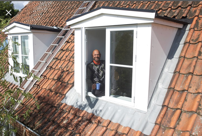 Man ler i ett nybyggt fönsterdormer på ett tegeltak, håller en kopp.