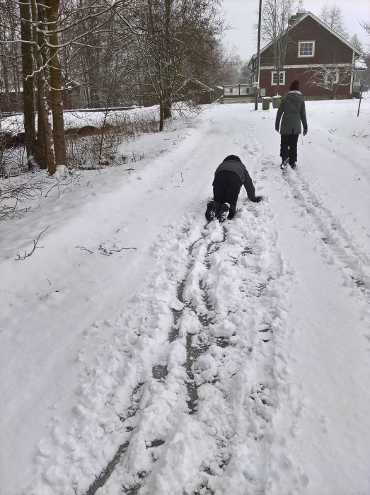 Två personer på en snöig väg nära ett hus, en böjer sig ner för att flytta snö.