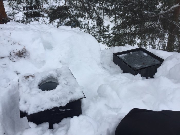 En öppning i snön på ett tak bredvid en frånluftsfläkt, omgiven av träd.