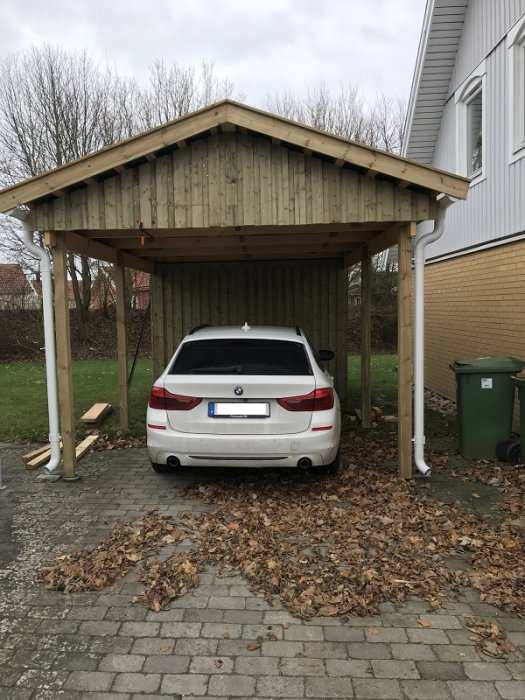 En vit bil parkerad under en nybyggd carport med träpanel och löv på marken.