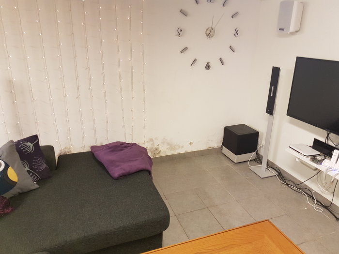 Källarrum med synlig fuktskada på väggen bakom en soffa och vid golvkanten, modern inredning med ljusslingor och klocka.