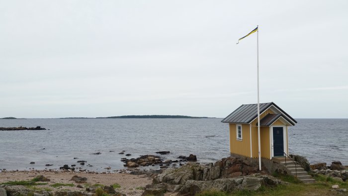 Gult litet hus vid havet i Torekov med svensk flagga på en flaggstång.