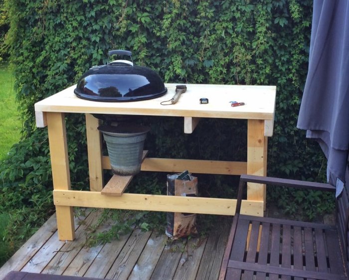 Hembyggt grillbord i trä med svart klotgrill ovanpå, placerat på en träterrass vid häck.