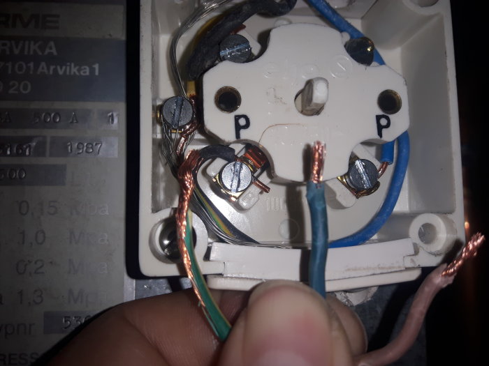 En hand håller tre avskalade kablar nära en öppen strömbrytare med anslutna ledningar och märkningen "P".