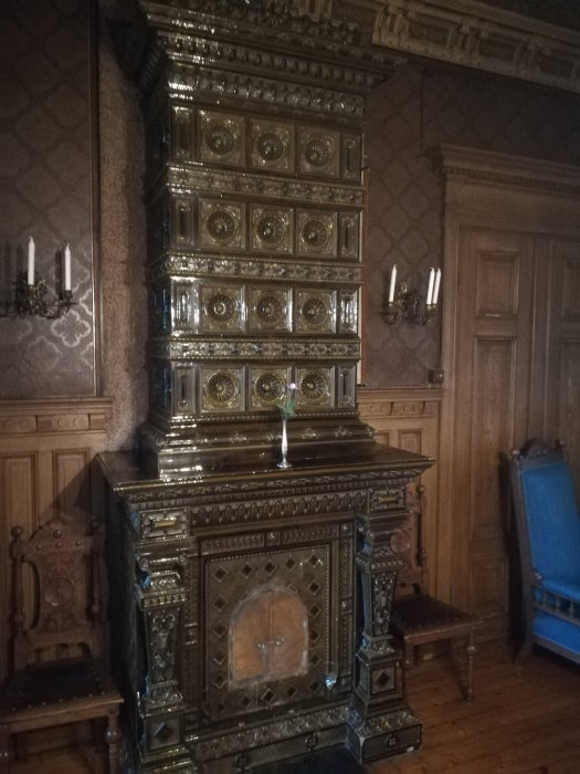 Ornate antique tiled stove in a room with patterned wallpaper and wooden details, hinting at ceiling issues.