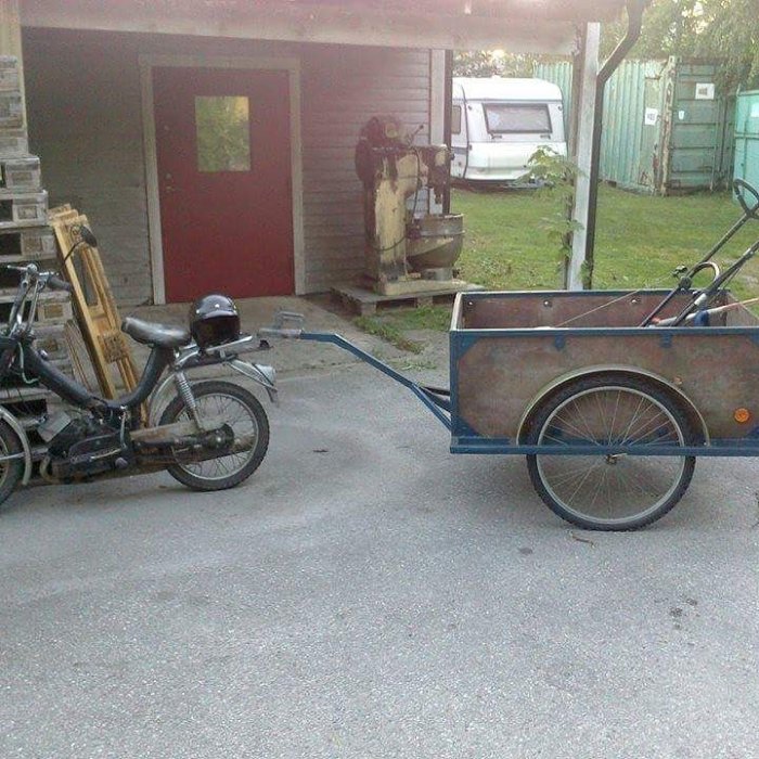 Moped med släpvagn parkerad framför en byggnad, använd för att transportera olika föremål.