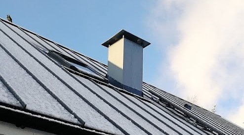 Snöbelagt tak med en stor spisfläkt och en mindre avloppsventil bakom en stege; klar himmel i bakgrunden.