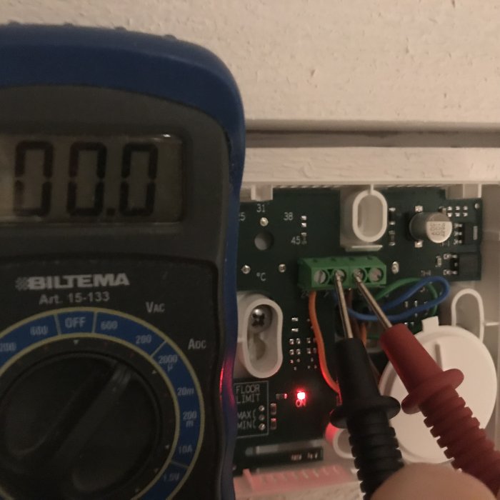 Multimeter visar 0.00V bredvid en termostat med tänd röd lampa och elektriska ledningar.