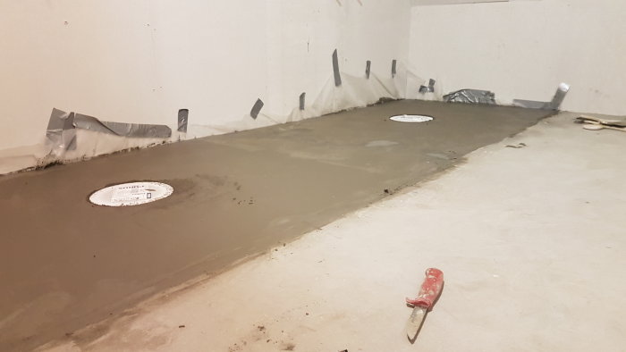 Rå betonggolv med två synliga avlopp och tätningstejp längs väggen i ett pågående badrumsprojekt.