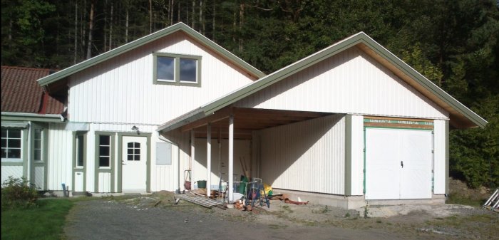 Grått hus från 90-talet med integrerad carport och garage med lutande tak på krypgrund, inbyggd halv carport och skog i bakgrunden.