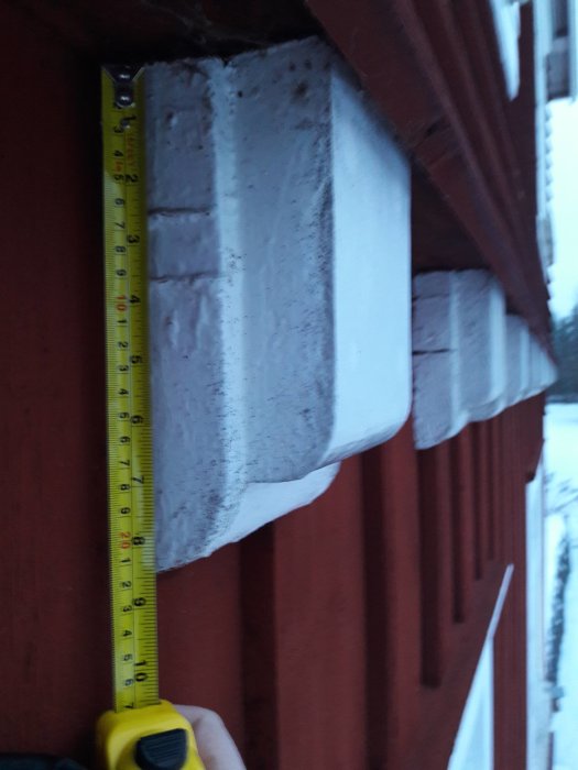 Mätning av bjälke på en vind med måttband som visar dimensionerna, vid sidan av en röd trävägg.