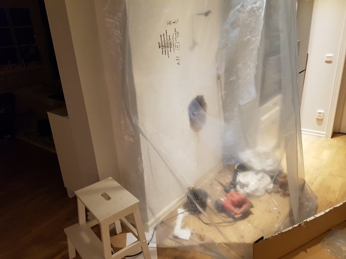 Renoveringsplats med plastskydd, vit pall och byggverktyg, ingen källaröppning synlig.
