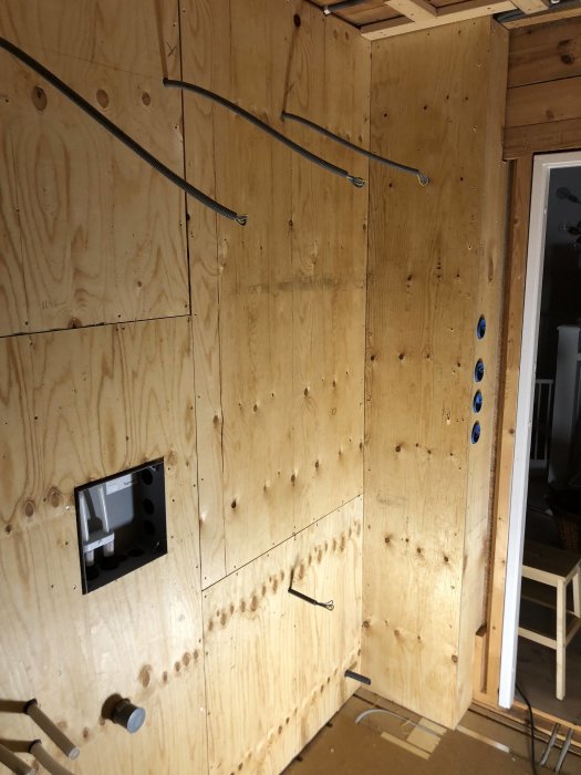 Delvis färdigställd badrumsvägg med oslipad plywood med utskärningar för installationer och synliga elkablar.