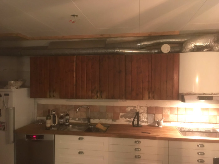 Ett kök under renovering med nya underskåp och ett träpanel bakom synliga ventilationrör och en plåtlåda på väggen.