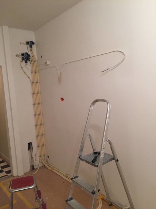 Översiktsbild av en vägg under renovering med elinstallationer och rör, samt en stege i förgrunden.