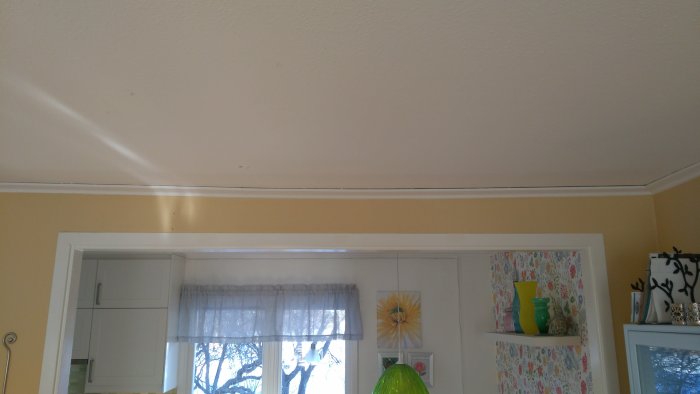 Kuvassa näkyy kallistunut katto-osa olohuoneessa, aukko johtaa keittiöön ja taustalla on ikkuna verhoilla.