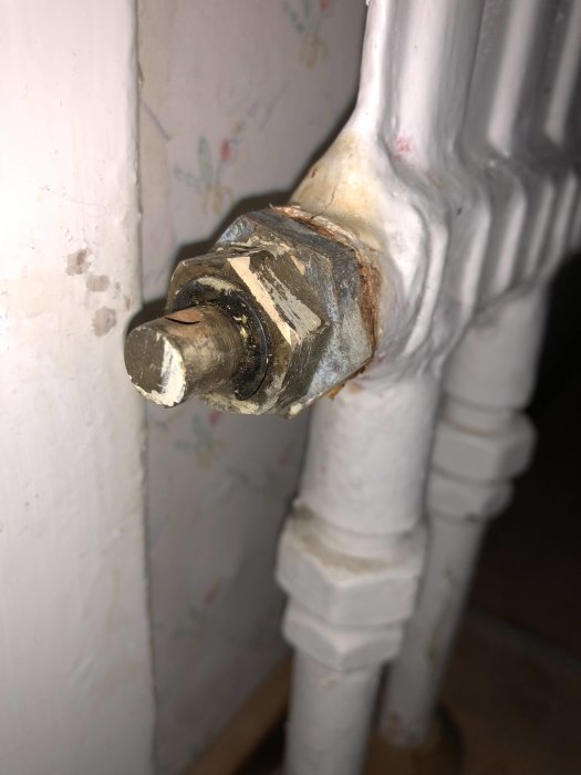 Närbild på en vitmålad gammal radiatorventil med slitage och korrosion mot en tapetserad vägg.