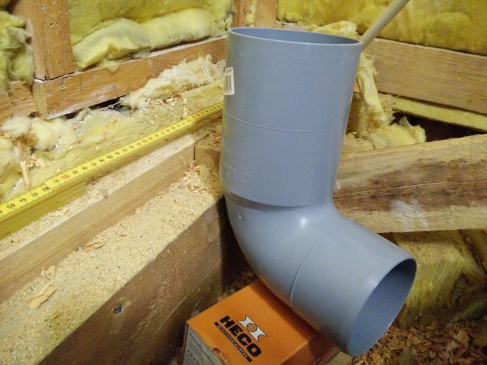 Harmaa muovinen viemäriputken kulmakappale kallistettuna puukehikon ja eristeen vieressä; vieressä mittanauha.