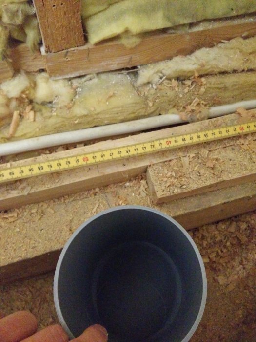 Käden pitelemä putki, eristeet ja mittanauha rakennustyömaalla seinän viereisessä tilassa.