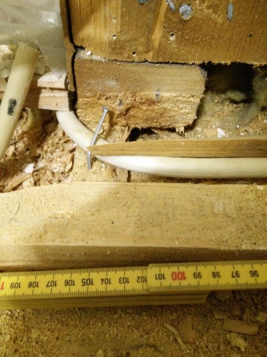 Lähikuva puulevyistä ja putkista, mittanauha puualustan päällä huoneen rakenteissa. Kynsistä ja puunpölystä näkyvissä yksityiskohtia.