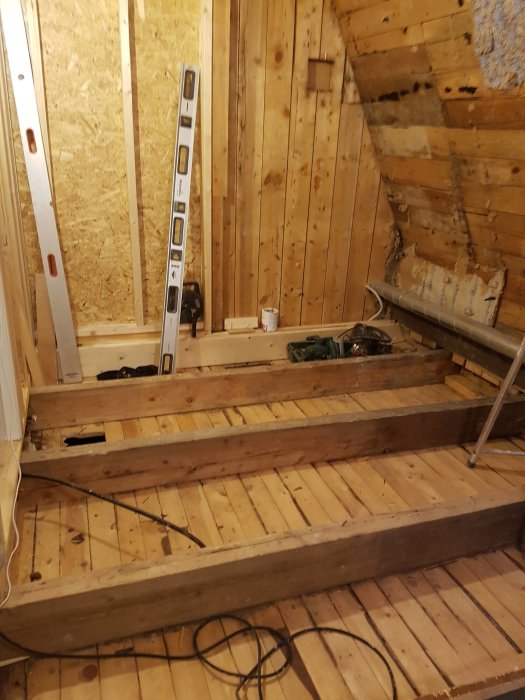 Vanhaa puurakenteista seinää ja portaita, joissa puupalkit lepäävät lattialaudalla. Rakennustyövälineitä näkyy taustalla.