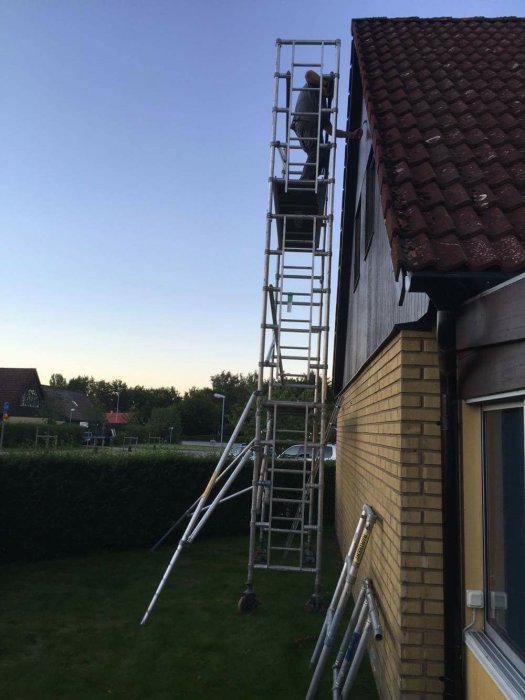 Person som målar hög del av hus med hjälp av uppbyggd ställning vid sensommar.