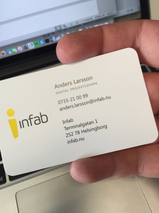 Hand håller ett visitkort från Infab med namnet Anders Larsson och titeln Digital Projektledare framför en datorskärm.