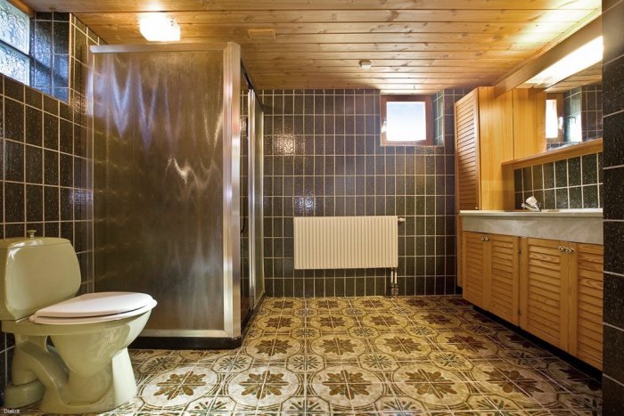 Gammalt badrum med mörkt kakel, frostade glasduschväggar, retro mönstrat golv och träpaneltak före renovering.
