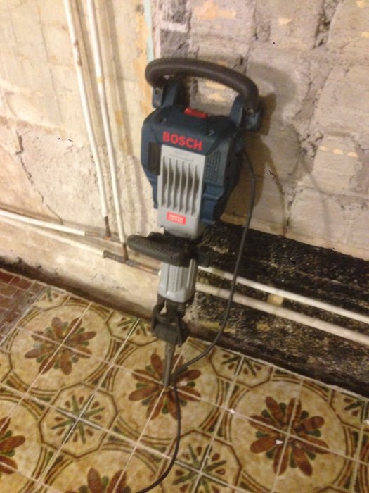 En Bosch borrhammare står på ett mönstrat källargolv med synliga rör för golvvärme och avskalad vägg.