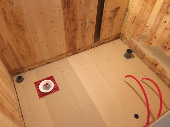 Renoverat badrumsgolv med nyinstallerat 22 mm golvspån, röda rör syns och avloppsrörinstallering.