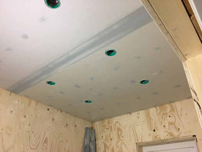 Tak med isolering och utsågade hål för spotlights ovanför plywoodvägg under renovering.