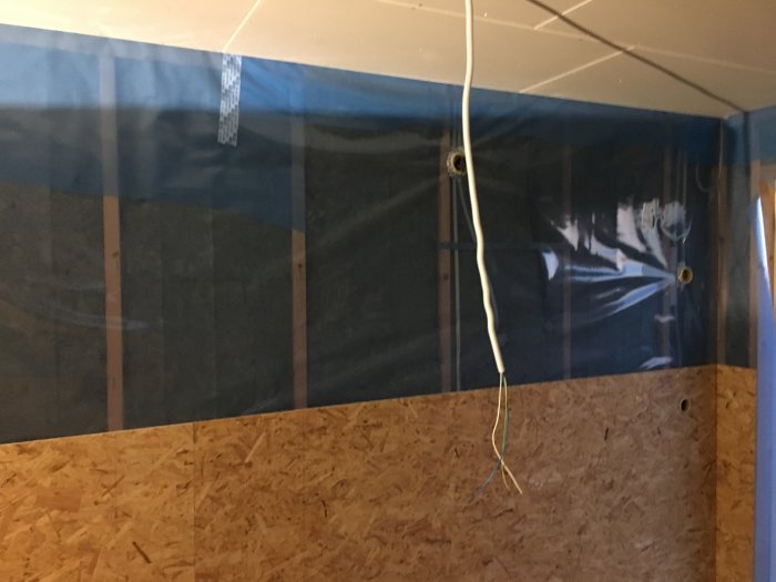 Hörn av ett rum under renovering med OSB-skivor installerade på nedre delen av väggen och plastskydd ovanför.