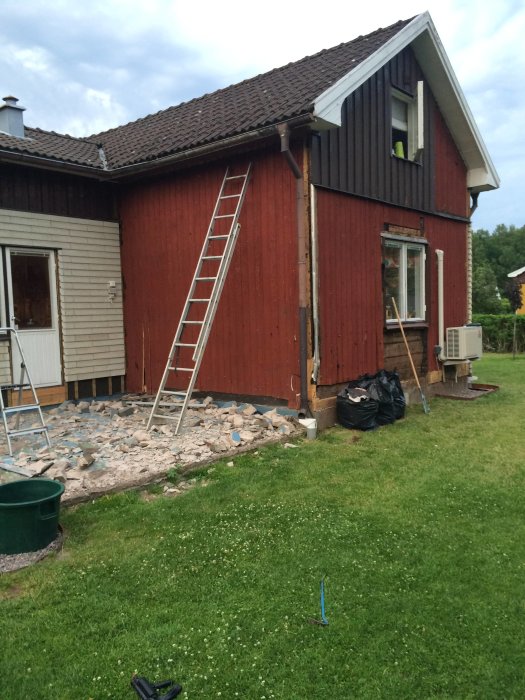 Röd träfasad på hus med rivet eternittak och byggnadsställning, rivningsmaterial på marken.