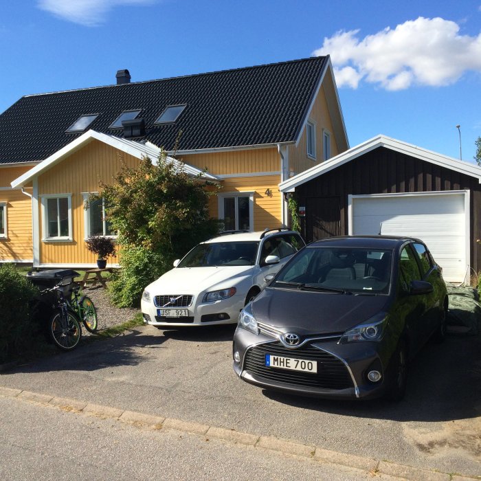 Gult hus med svart tak och garage, två bilar framför, och en ettåring som står i gruset.