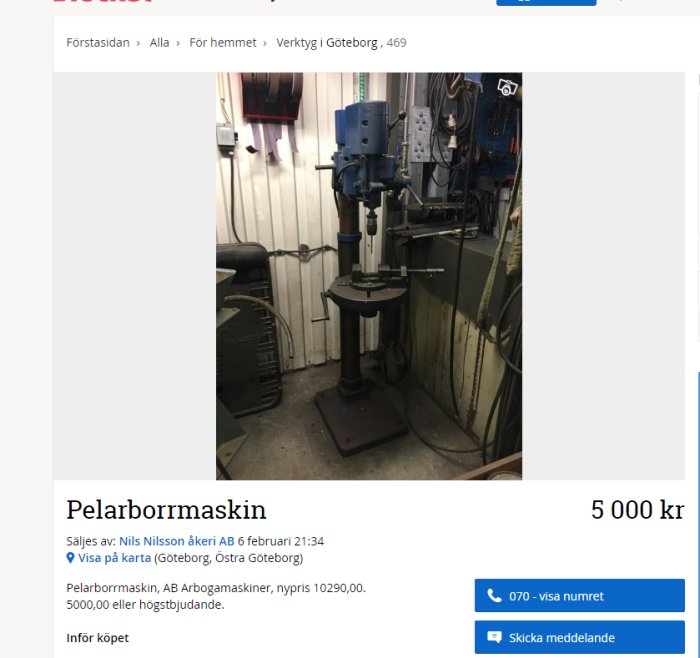 Pelarborrmaskin i ett verkstadsutrymme med verktyg och kablar på väggen i bakgrunden.