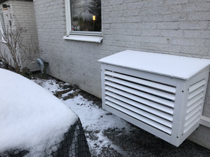 Husets vitmålade tegelvägg med fönster och en täckt ventilationsrör, markerad potentiell grävplats för garage.