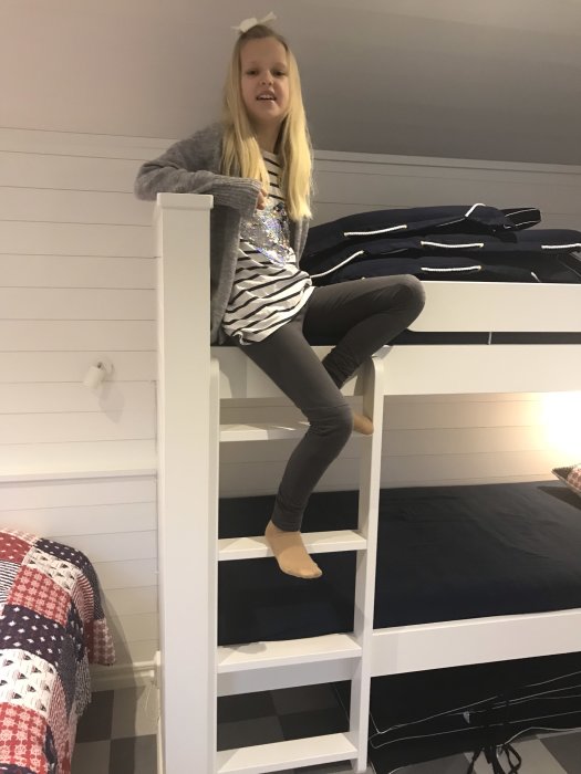 En flicka sitter på en vit loftsäng i ask med många trappsteg, intill en säng med blått överkast.