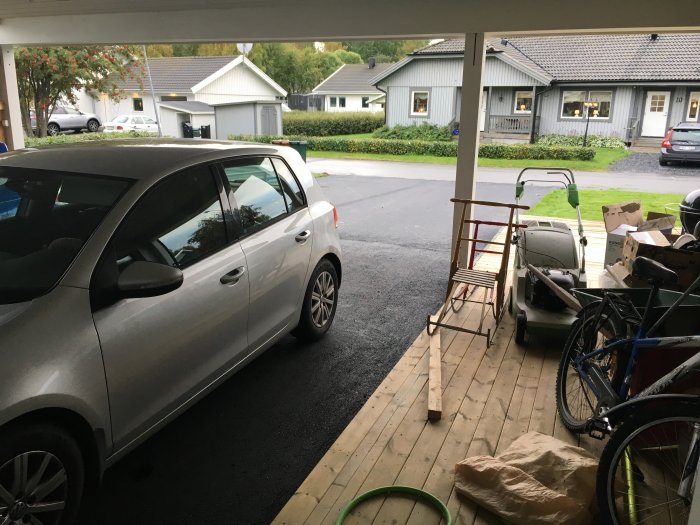 Vy från en carport med en parkerad bil, byggmaterial och verktyg, mot ett bostadsområde.