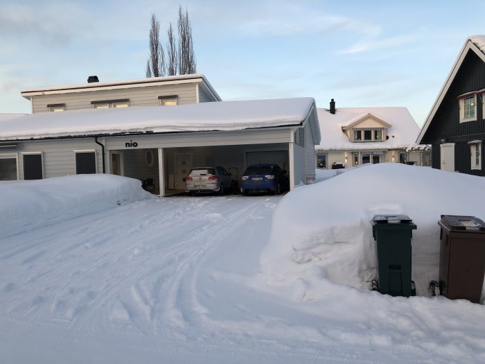 Vinteruppfart med bilar parkerade utanför ett vitmålat hus, snötäckt landskap och sopkärl.